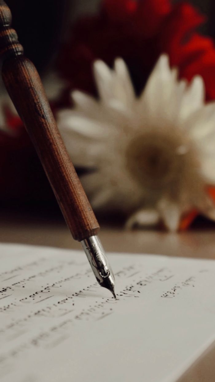 About Close-up of a wooden fountain pen writing calligraphy on paper with blurred flowers in the background.