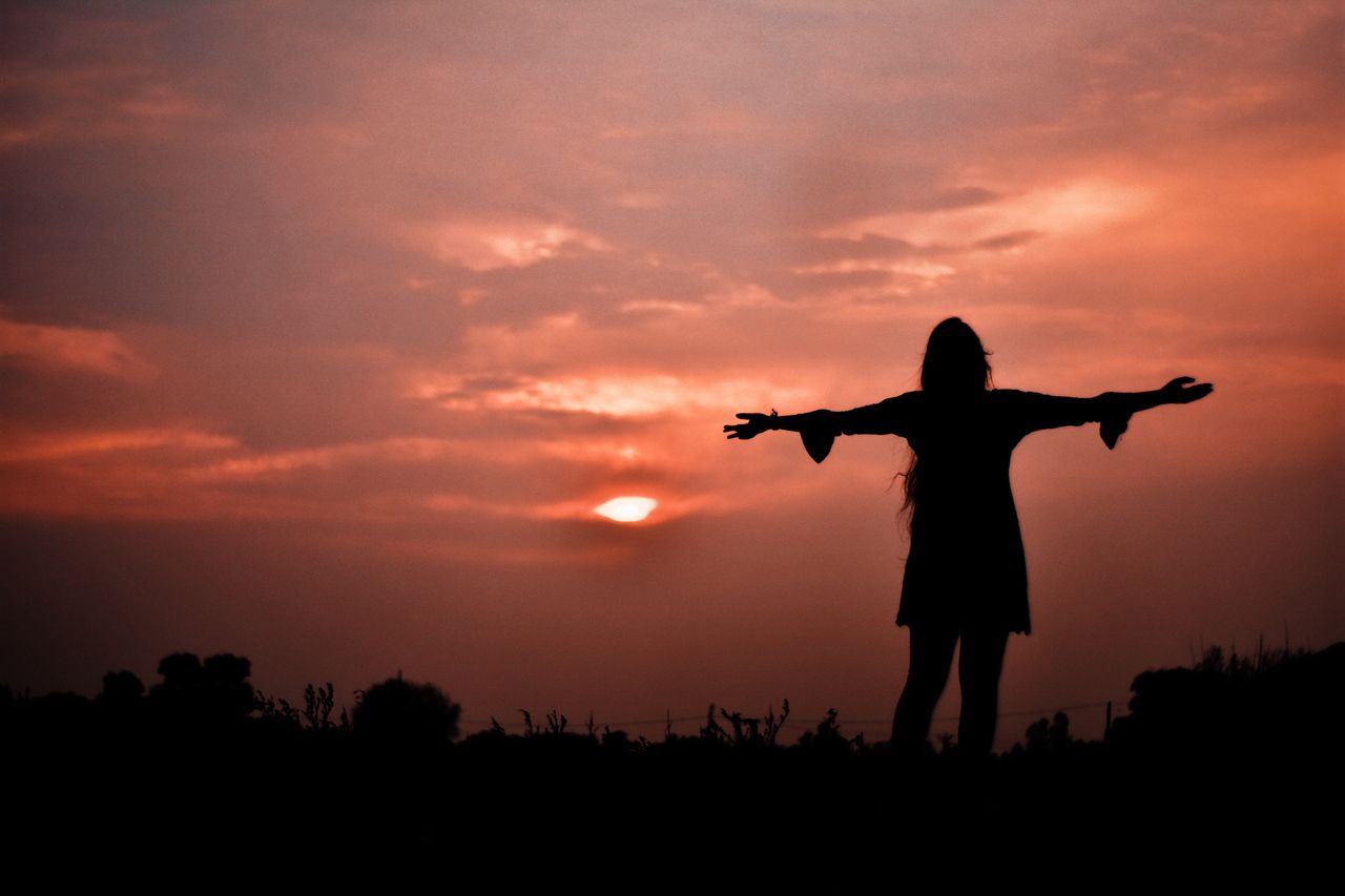 About A woman stands in silhouette with arms open, embracing a dramatic sunset in India.
